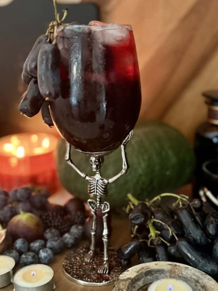 Gothic-style skeleton wine glass holder with dark red wine against a cozy, autumnal background featuring a watermelon, grapes, and candles. Perfect for Halloween or themed wine nights.