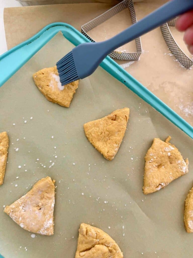 Unbaked pumpkin scones being brushed with milk on a lined baking sheet.