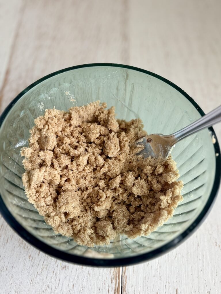 Crumbled streusel topping in a clear glass bowl with a fork on a wooden surface, perfect for baking or dessert recipes.
