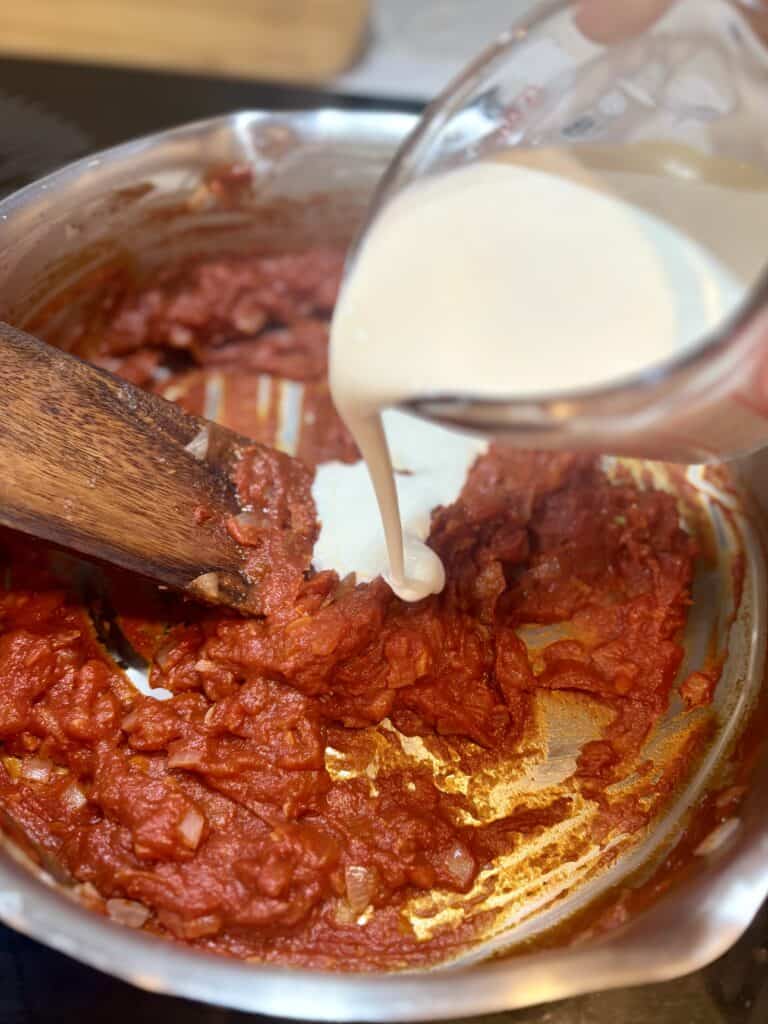 Cream pouring into simmered tomato sauce in a stainless steel pot, with a wooden spoon stirring, preparing a homemade pasta or pizza sauce.