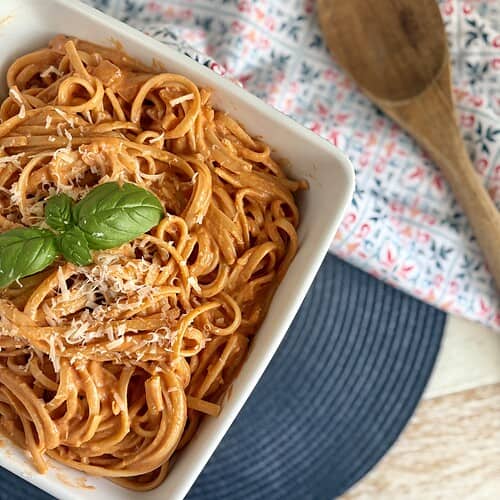 Creamy tomato pasta with shredded cheese and fresh basil leaves in a white dish, served on a navy placemat with a wooden spoon and patterned cloth. Perfect for quick and delicious homemade Italian recipes.