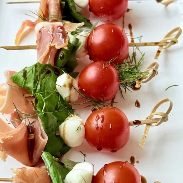 Fresh tomato, mozzarella, basil, and prosciutto skewers drizzled with balsamic glaze on white background.