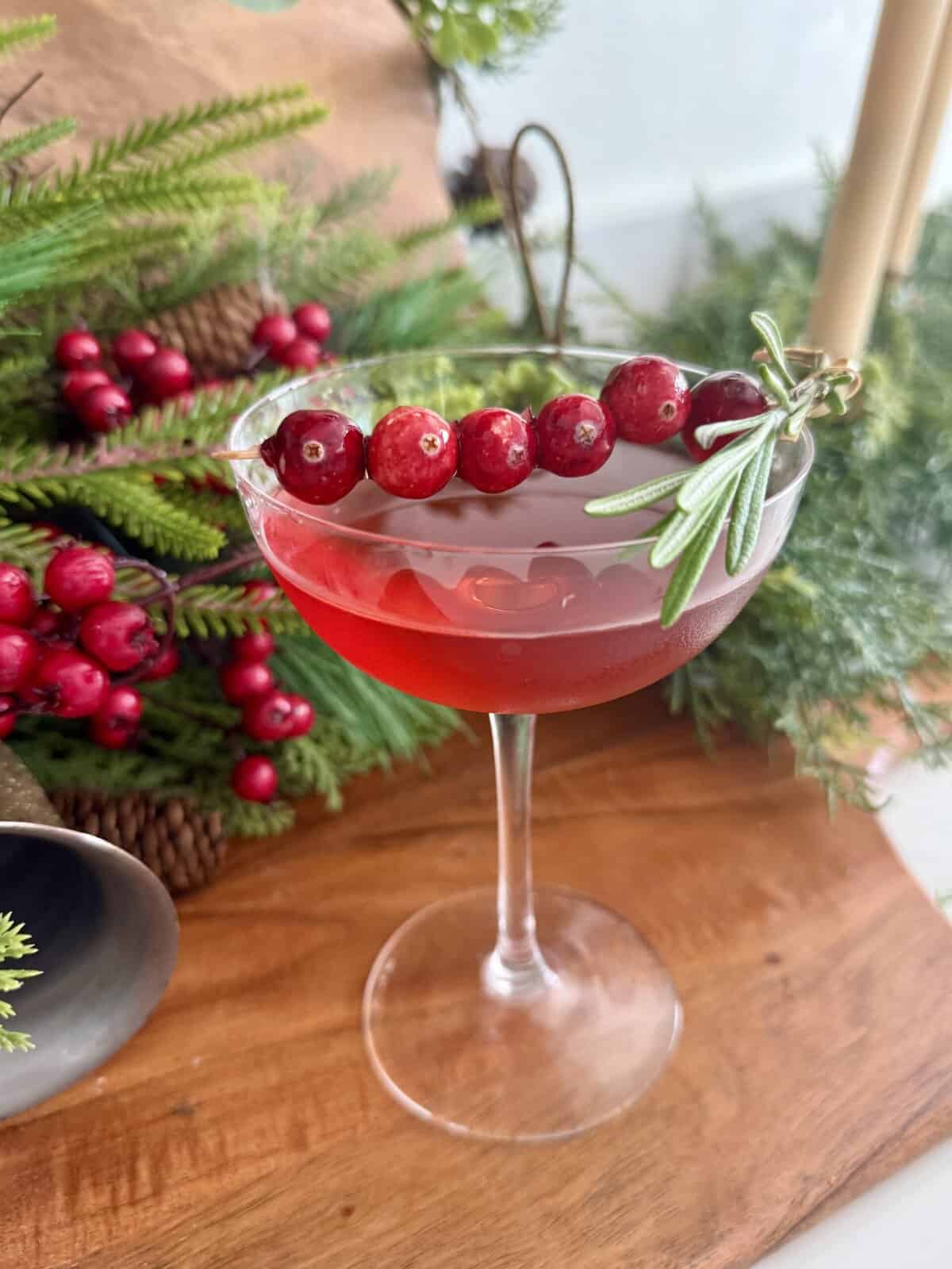A festive red berry cocktail garnished with fresh rosemary, served in an elegant coupe glass, ideal for holiday celebrations. The background features holiday greenery and natural wood, creating a cozy and festive atmosphere perfect for winter gatherings and Christmas parties. The drink's vibrant color and garnishes make it a beautiful addition to your holiday table or special occasion.