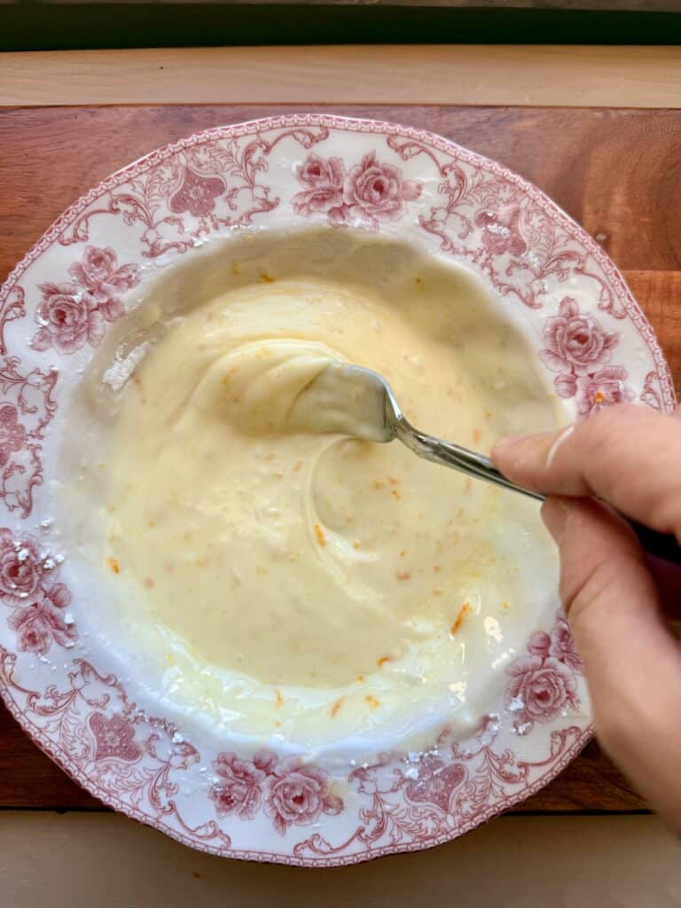 Mixing together powdered sugar, orange zestt, and orange juice in a white and red plate on a cutting board with a silver fork to make an icing