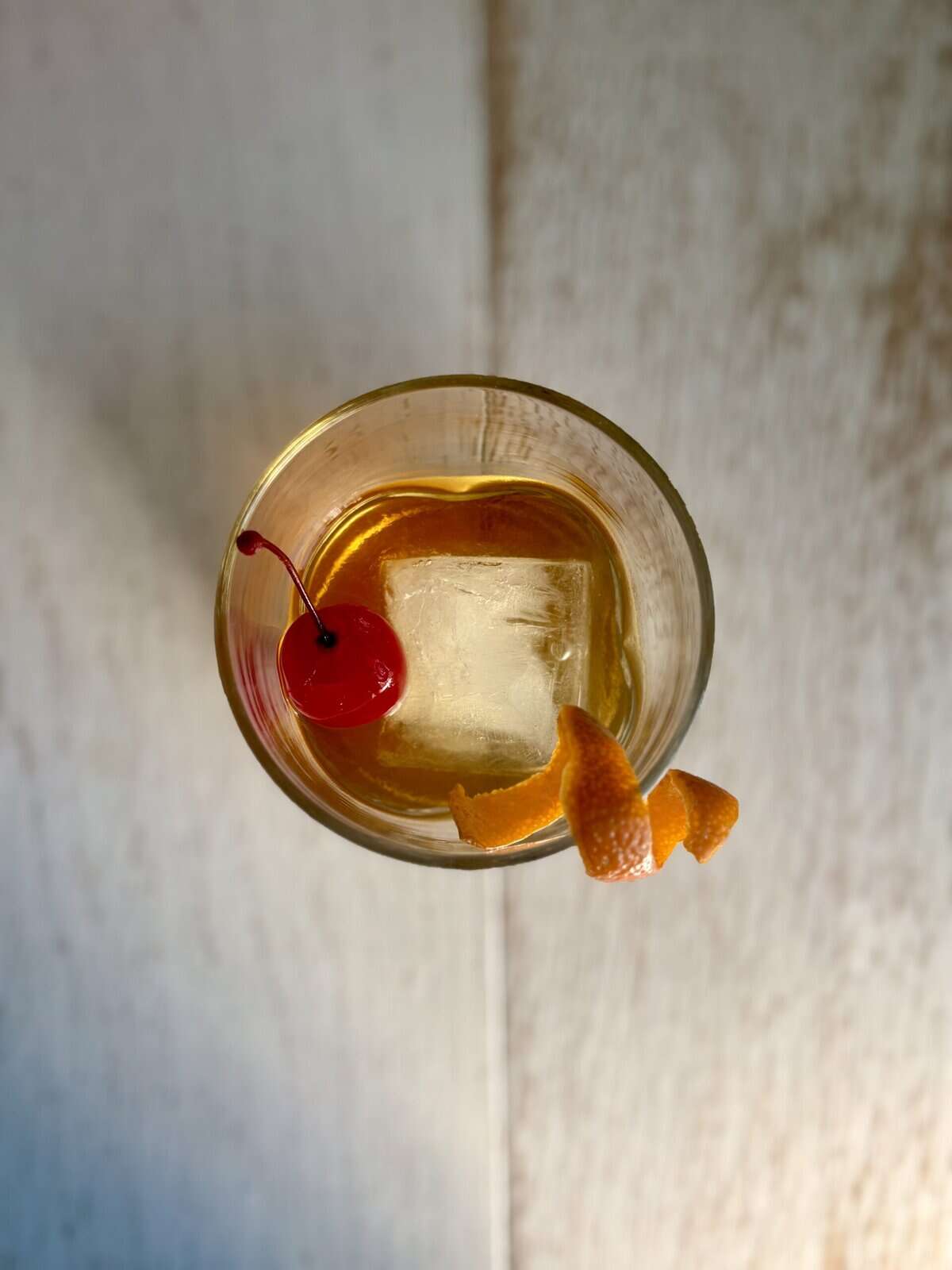 Bright cocktail with a cherry and orange peel garnished on a clear glass with ice, viewed from above on a rustic wooden surface.