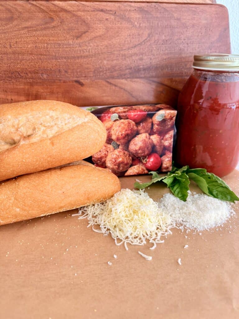 Jar of tomato sauce, box of frozen turkey meatballs, tow sub rolls, shredded mozzarella cheese and grated Parmesan cheese, and basil leaves on brown parchment paper.