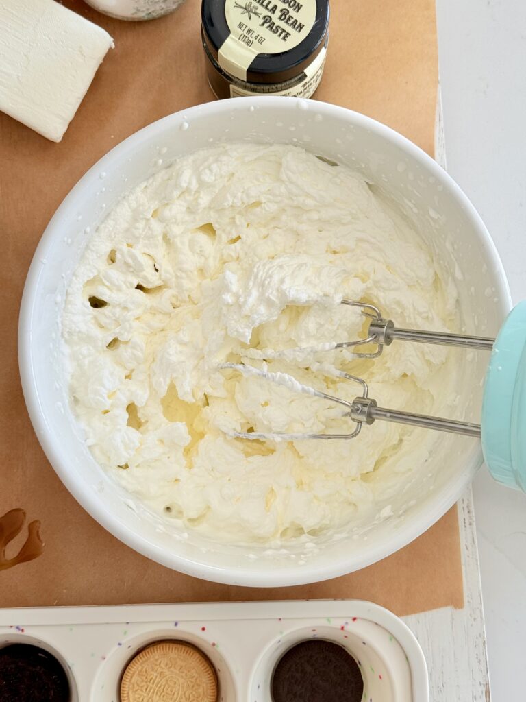 An electric blue hand mixer sits in a white bowl of whipped heavy cream on brown parchment paper. Below it is a silicone muffin tin with Oreos at the bottom of each cup, and a container of vanilla bean paste rests on top of the bowl.