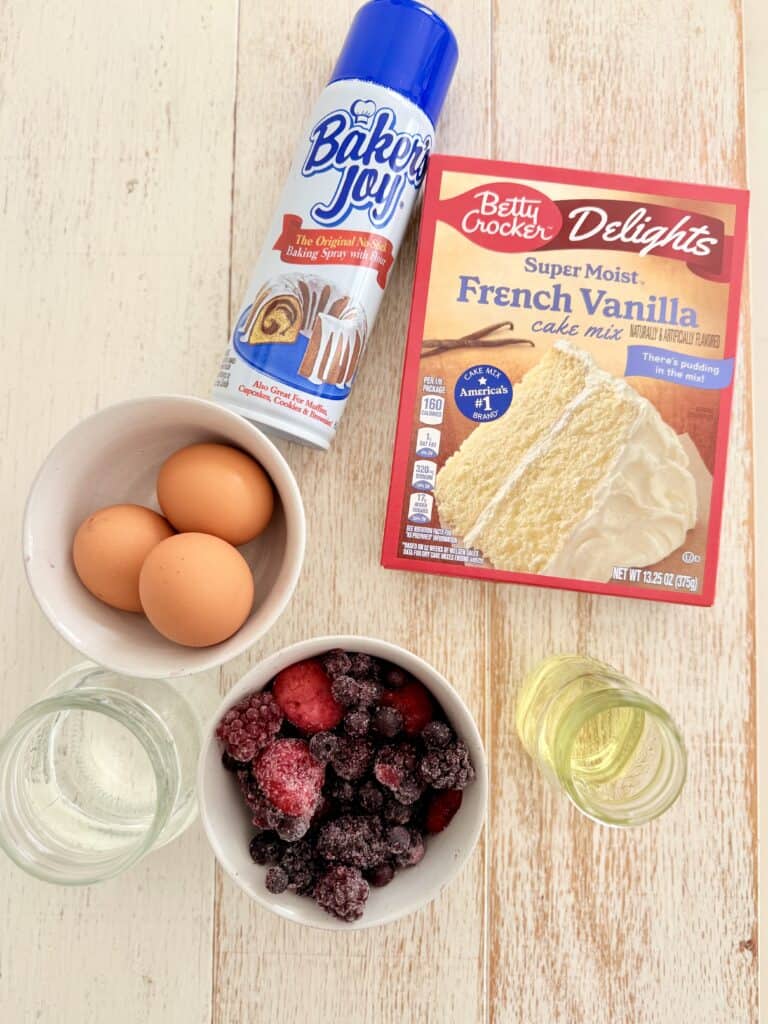 Baking spray, Betty Crocker French cake mix, three brown eggs, and a white bowl, water and a clear glass jar, frozen berries in a small white bowl, and vegetable oil in a small glass jar, all on the white countertop.