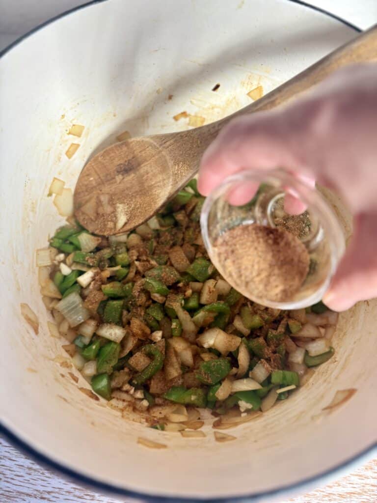 Sautéed onions and green peppers in a pot bowl, with spices being added for a flavorful homemade dish, showcasing fresh ingredients and cooking process.