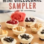 Cream cheese mini cheesecakes topped with Oreo cookies, graham crackers, and fruit preserves, arranged on a wooden surface with a pink rose in the background, promoting homemade mini cheesecake recipe.