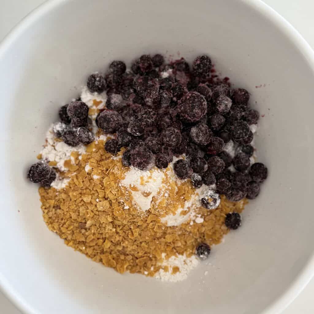 Crushed cornflakes, Bisquick and frozen berries blueberries in a white mixing bowl on the counter