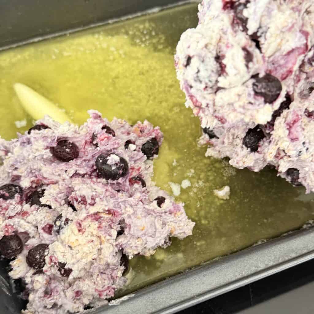 Melted unsalted butter in an 8 x 8 baking dish with the biscuit, corn flake milk, frozen blueberry dough spread out on top on top of the stove top
