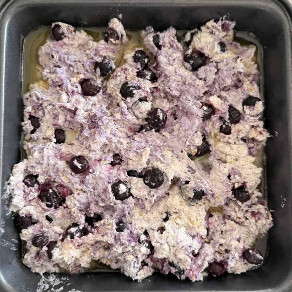 Melted unsalted butter in an 8 x 8 baking dish with the biscuit, corn flake milk, frozen blueberry dough spread out on top on top of the stove top
