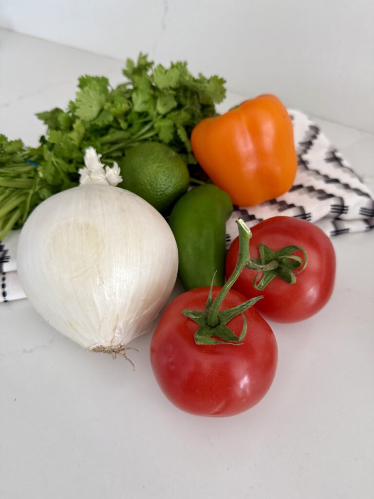 On the white counter or two ripe tomatoes that are red jalapeño lime, a large, white onion and orange, bell pepper and cilantro on top of a hand towel that is white, black and gray striped