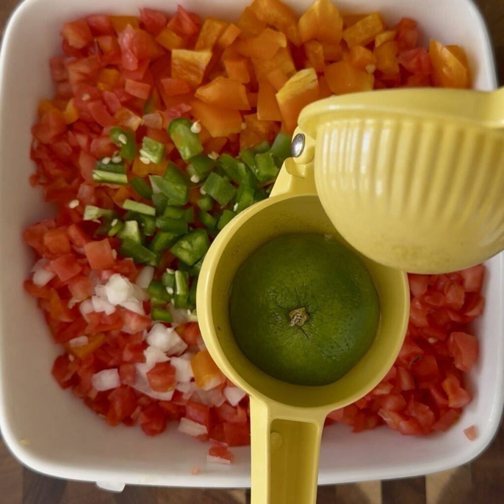 This vibrant pico de gallo features a colorful mix of chopped tomatoes, onions, jalapeños, and cilantro, offering a fresh and zesty flavor. Ideal for enhancing Mexican dishes or serving as a healthy snack, this homemade salsa is a staple in Mexican cuisine. The image showcases a bowl of freshly prepared pico de gallo, emphasizing its bright colors and fresh ingredients, perfect for adding a burst of flavor to any meal. A yellow citrus extractor is a bout to extract juice from half a lime onto the pico de gallo ingredients on a white bowl on the counter