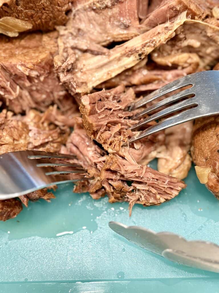 Juicy, tender pulled beef shredded with forks, perfect for sandwiches, served on a teal cutting board. Ideal for easy, flavorful meal prep and family dinners.