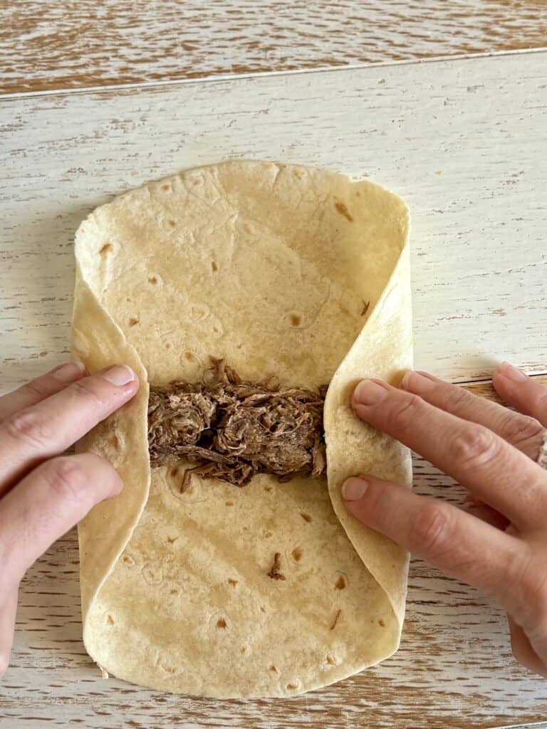 Hands folding a soft flour tortilla around seasoned shredded beef filling, ready to be rolled into a delicious homemade flauta on a rustic wooden surface.