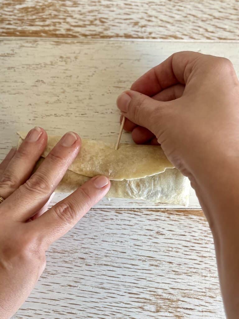 Hands sealing a flat with a toothpick on a rustic wooden surface, showcasing traditional homemade flautas preparation.
