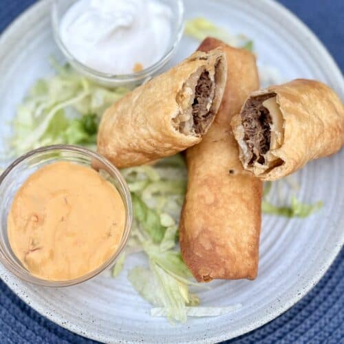 Crispy shredded beef flautas on a bed of lettuce next to bowls of sour cream and salsa con queso on a white plate.