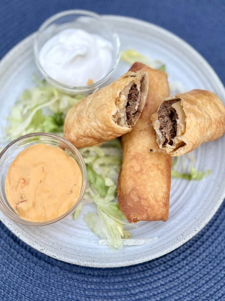 Crispy shredded beef flautas on a bed of lettuce next to bowls of sour cream and salsa con queso on a white plate.