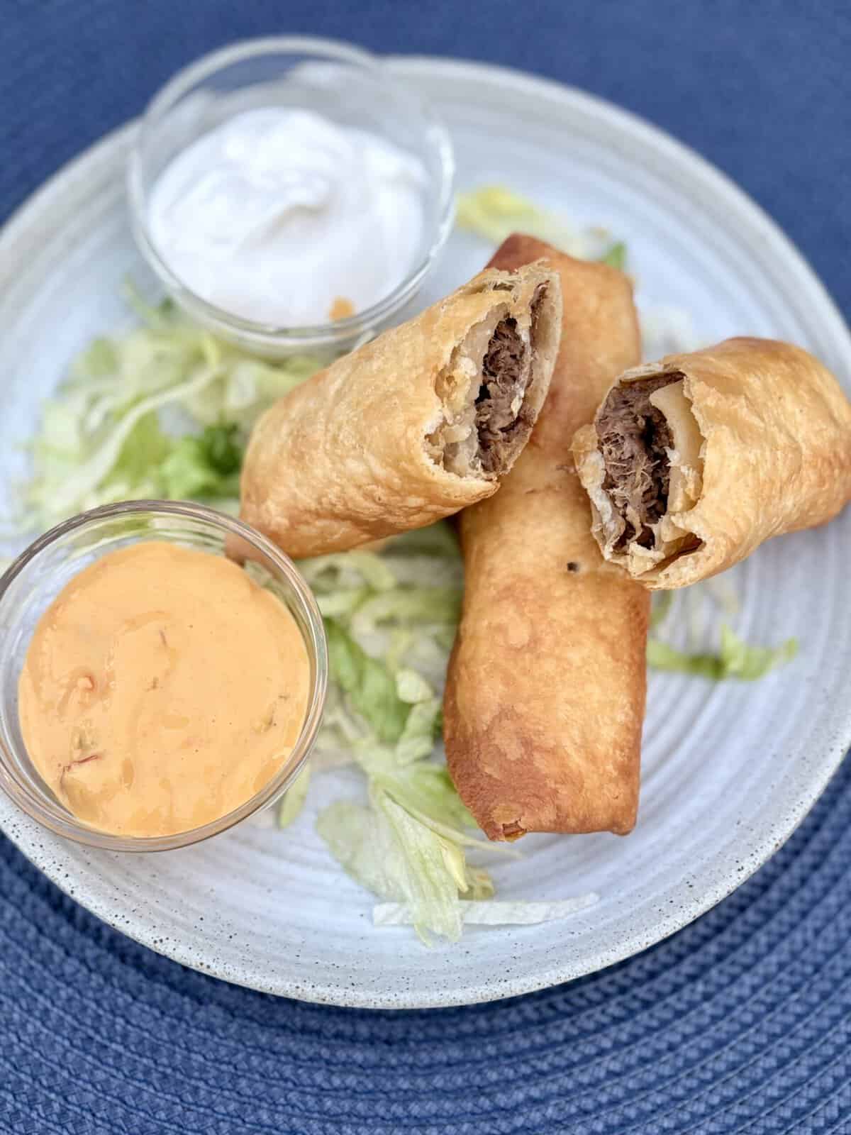 Crispy shredded beef flautas on a bed of lettuce next to bowls of sour cream and salsa con queso on a white plate.