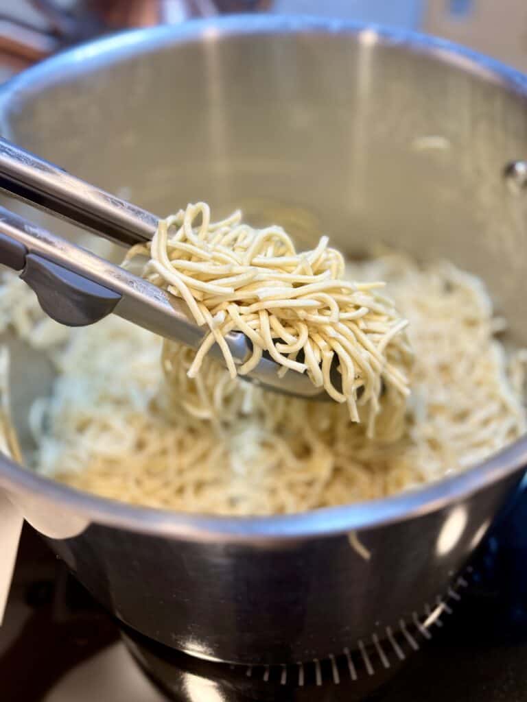 Fresh pancit canton noodles being lifted from a large stainless steel pot using tongs, showcasing a simple noodle dish.
