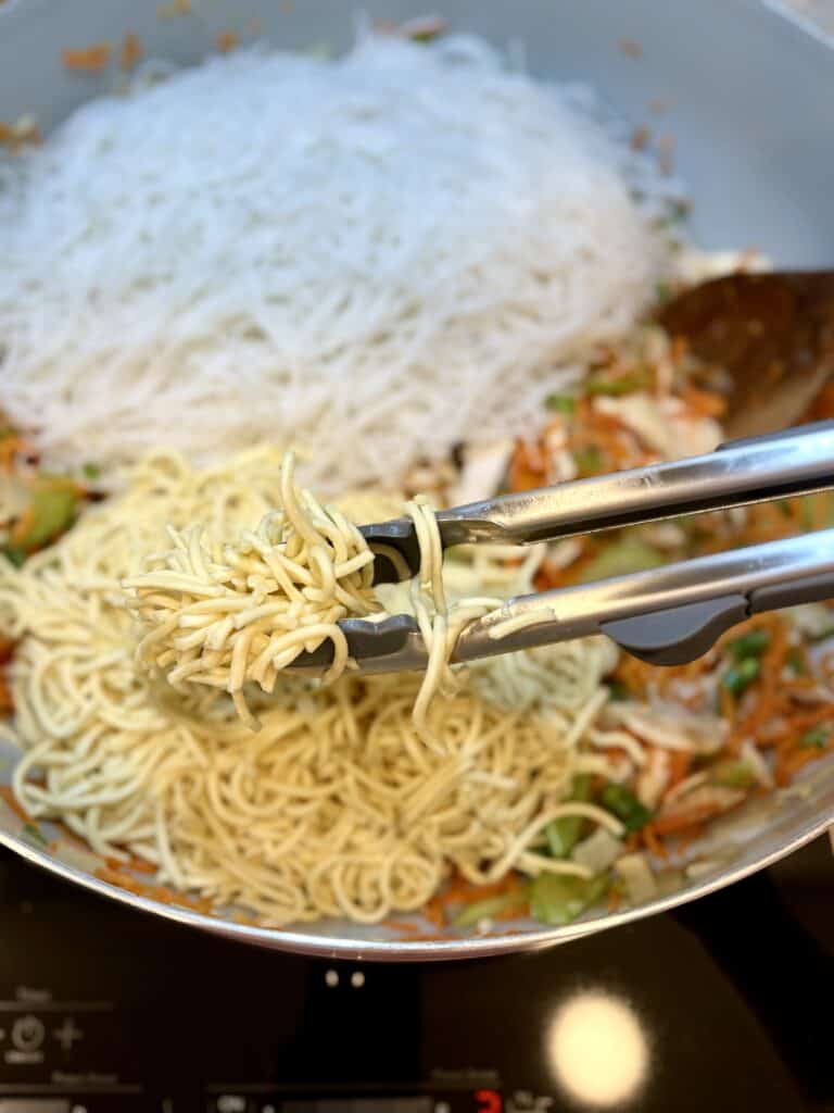 Tongs holding cooked yellow noodles over a skillet filled with stir-fried vegetables and rice stick, showcasing a colorful Asian-inspired dish ready to serve.
