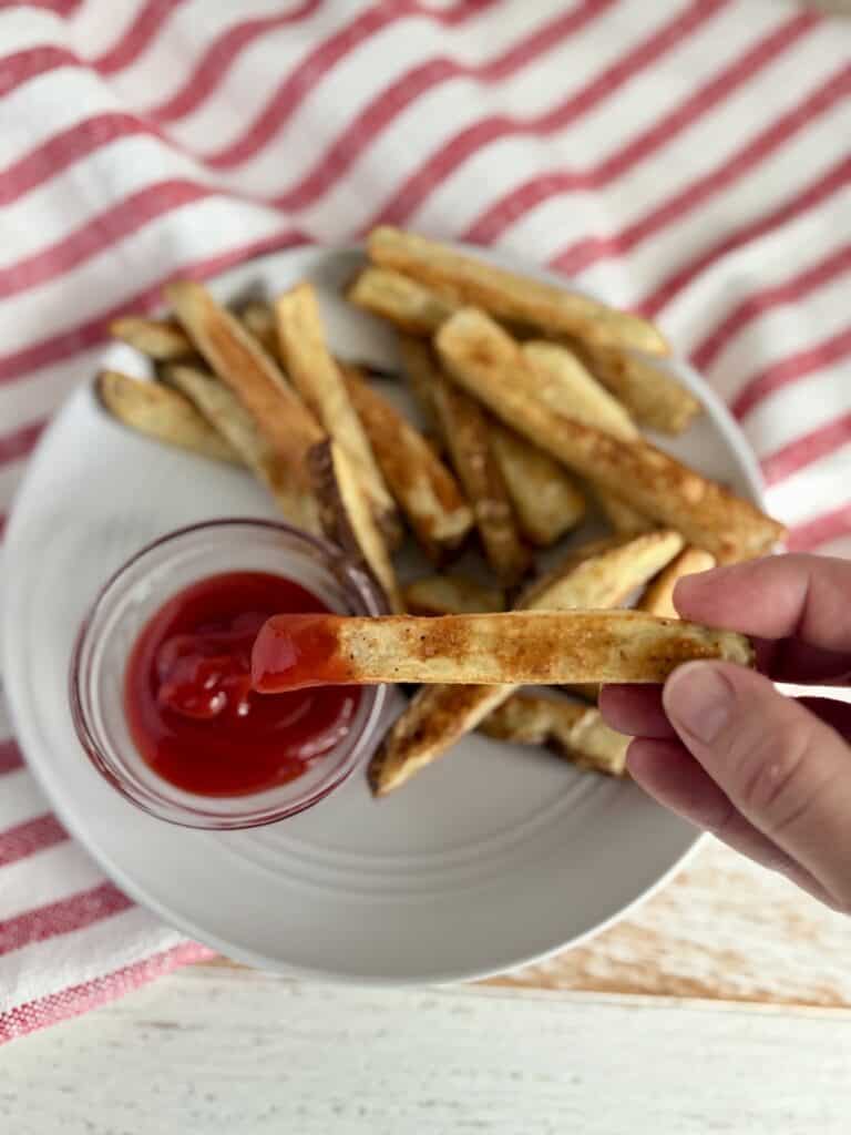 Golden crispy French fries served with a side of ketchup, perfect for snacking or sharing. Freshly cooked, seasoned potato fries on a white plate with a red and white striped cloth background.