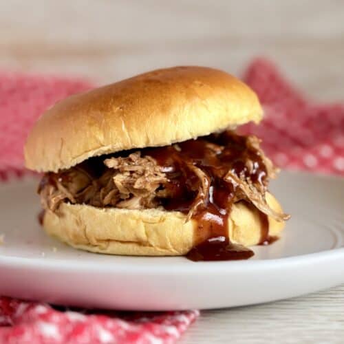 Juicy pulled pork sandwich with barbecue sauce in a soft bun, served on a white plate with a red checkered napkin in the background. Perfect for comfort food lovers.