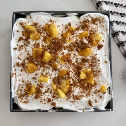 mango cheesecake royale dessert topped with whipped cream, crushed cookies, and mangoes in a black square dish.