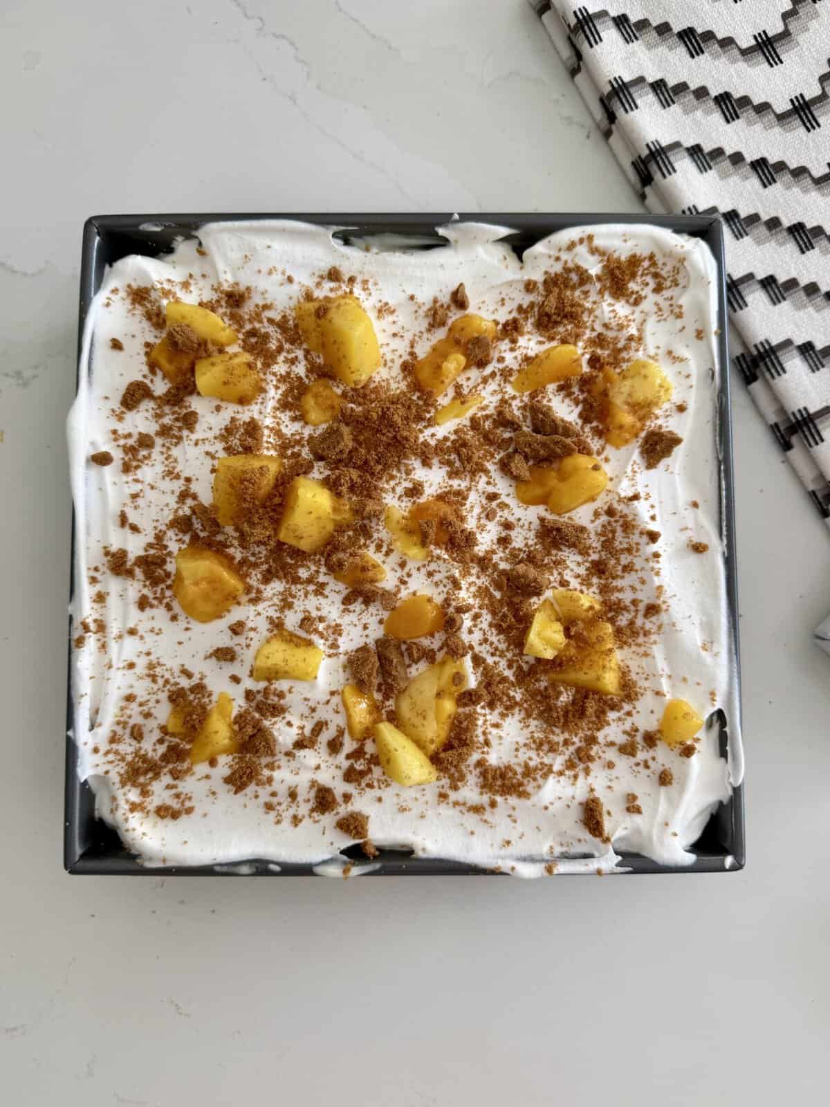 mango cheesecake royale dessert topped with whipped cream, crushed cookies, and mangoes in a black square dish.