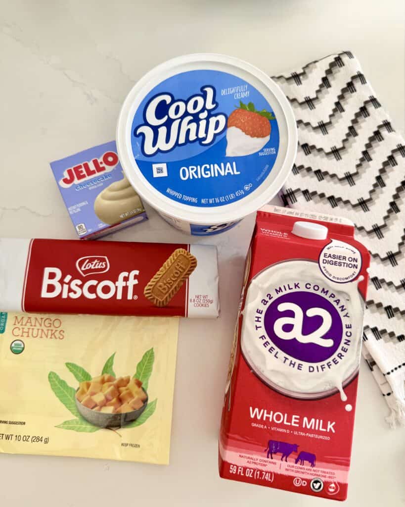 cool whip, cheesecake Jell-O instant pudding, whole milk, frozen mango chunks and Biscoff cookies with a black and white kitchen towel on the counter.
