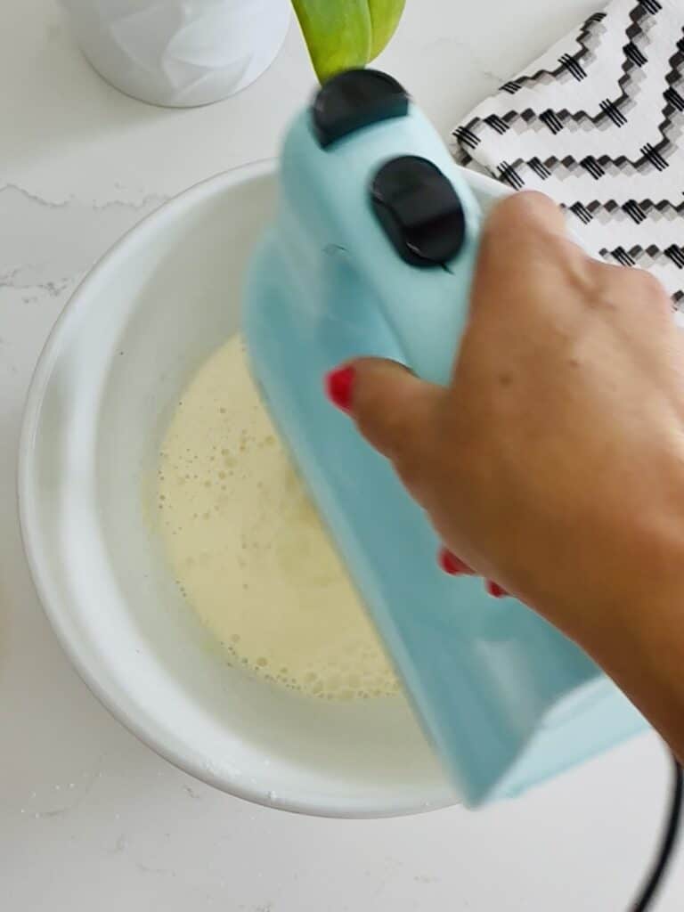 using an electric mixer to blend the milk and the instant cheesecake pudding all in a white bowl on the counter