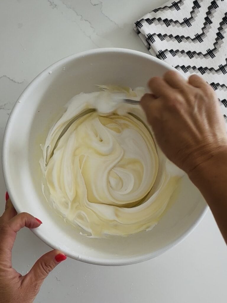 using a spoon to fold in Cool Whip into the instant cheesecake pudding. all in a white bowl on the counter