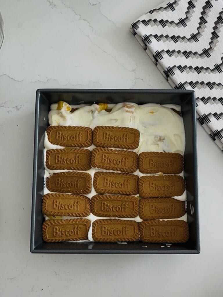 Delicious homemade layered dessert of mango and cheesecake topped with whipped cream, garnished with fresh mango pieces and a sprinkle of of biscoff cookies served in a black square dish for a sweet treat.