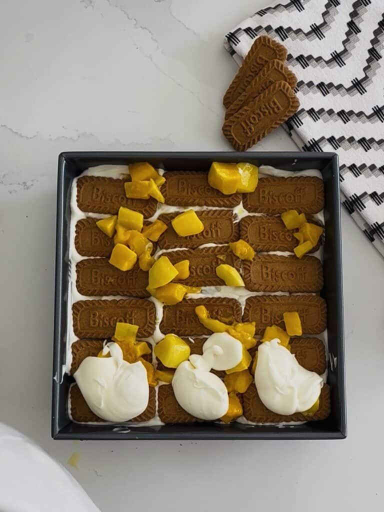 Delicious homemade layered dessert of mango and cheesecake topped with whipped cream, garnished with fresh mango pieces and a sprinkle of of biscoff cookies served in a black square dish for a sweet treat.