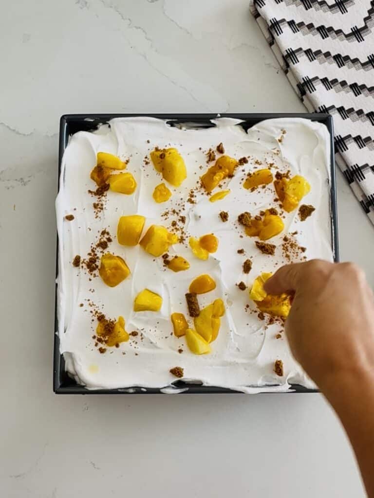Delicious homemade mango and cheesecake layered dessert with whipped cream, garnished with fresh mango pieces and a sprinkle of crushed cookies, served in a black square dish for a sweet treat.
