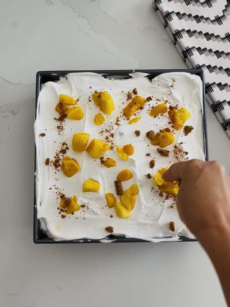 Delicious homemade mango and cheesecake layered dessert with whipped cream, garnished with fresh mango pieces and a sprinkle of crushed cookies, served in a black square dish for a sweet treat.