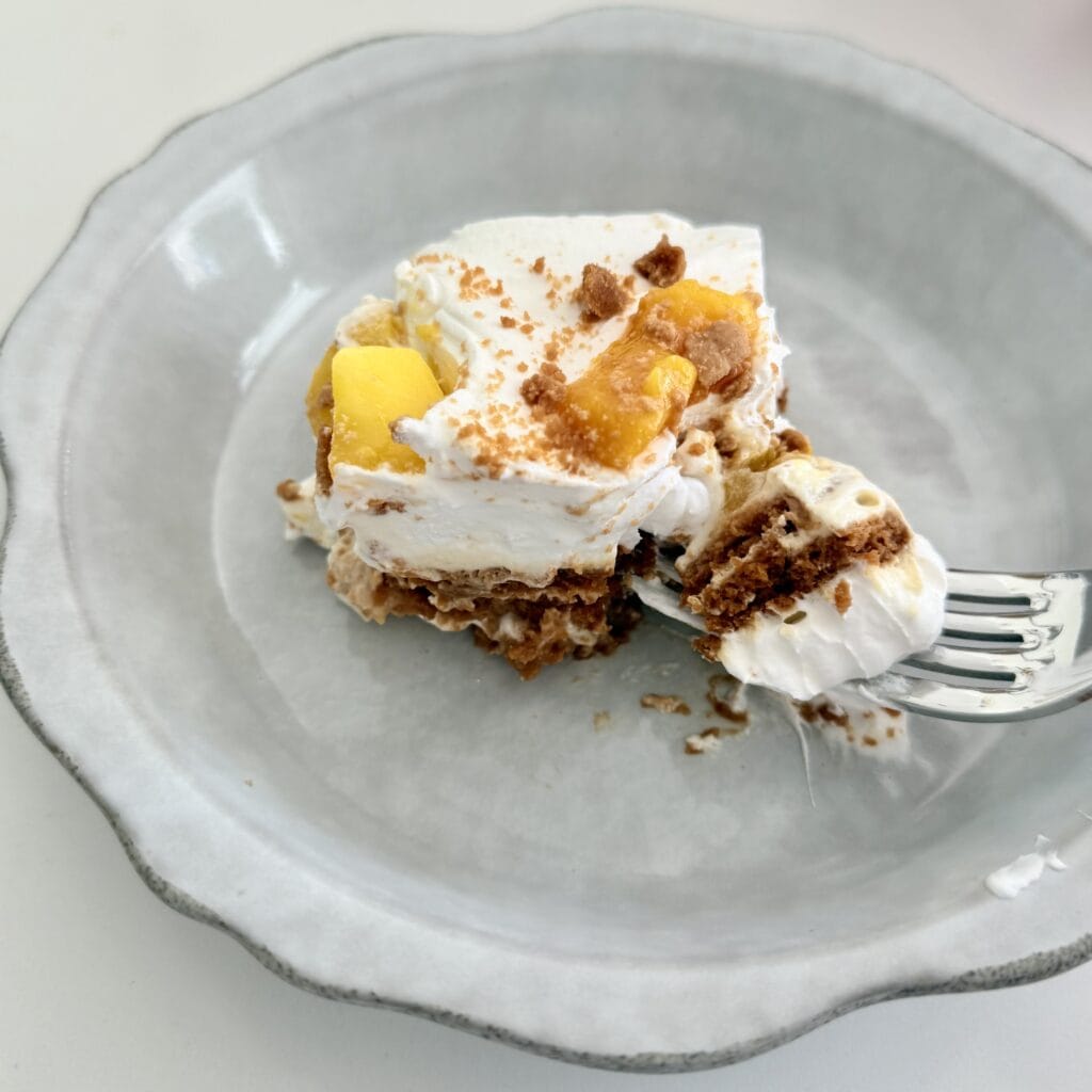 Delicious homemade mango and cheesecake layered dessert with whipped cream, garnished with fresh mango pieces and a sprinkle of crushed cookies, served in a black square dish for a sweet treat.