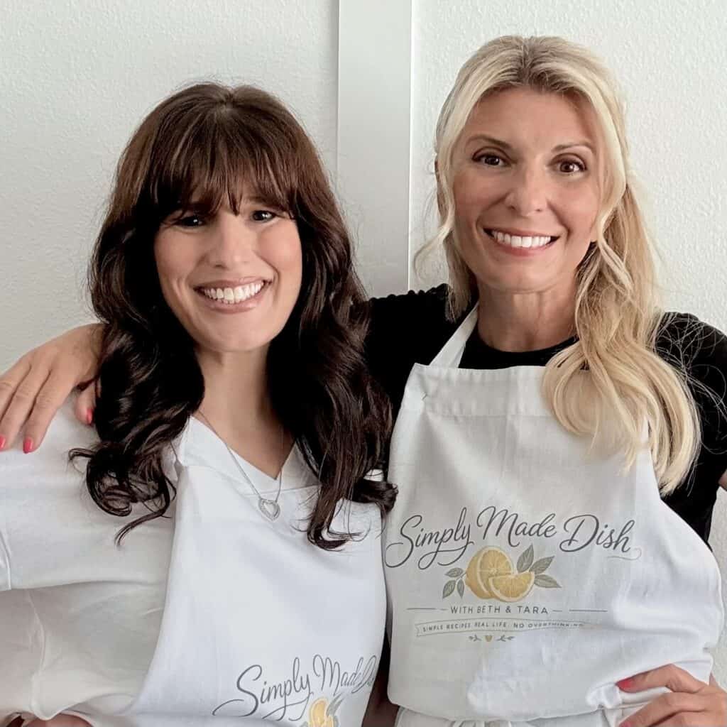 Two women smiling, wearing aprons with 'Simply Made Dish' logo, in a bright kitchen setting, promoting homemade cooking and culinary content.