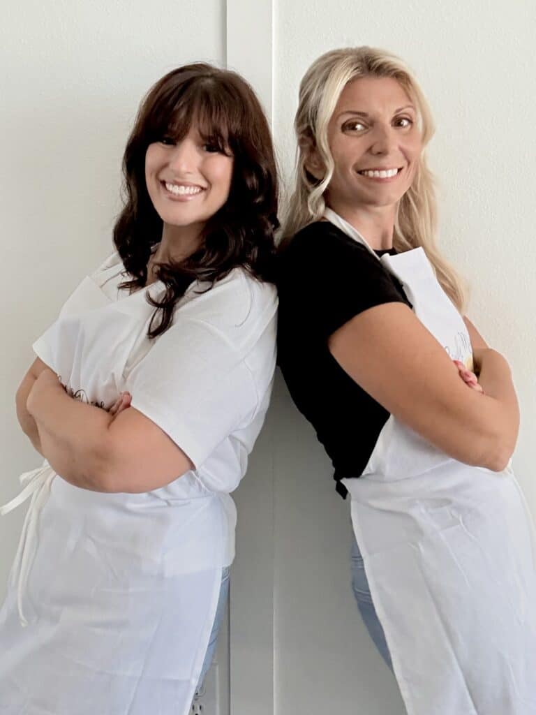 Two women, Beth and Tara, standing back-to-back with arms crossed, smiling, wearing white aprons, in a kitchen setting.
