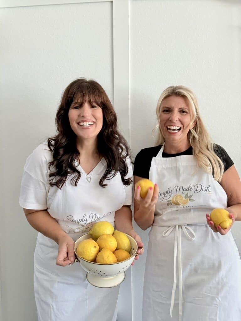 Two women in aprons holding lemons and a bowl of lemons, smiling in a bright kitchen setting.