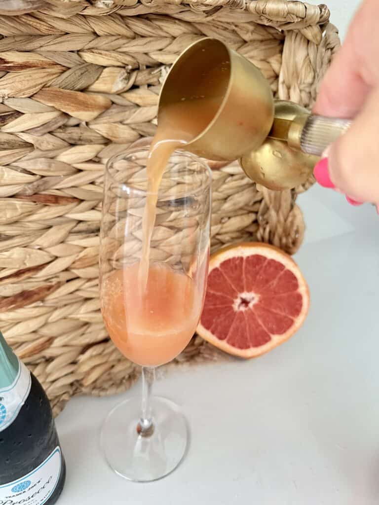 Pouring rub, red grapefruit juice into a champagne flute with a half slice of grapefruit on the kitchen counter behind it