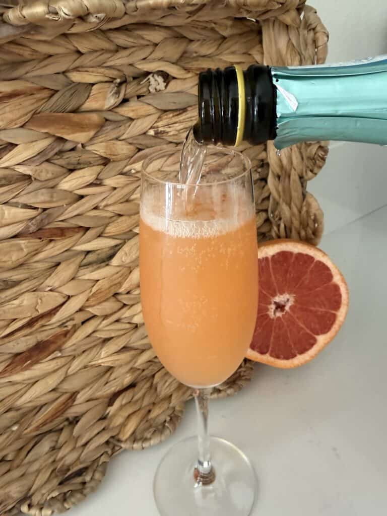 Pouring sparkling grapefruit beverage into a champagne flute with a halved grapefruit beside it, set against a woven basket background.