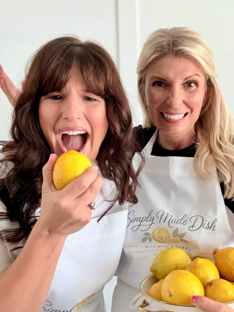 Two women in aprons smiling in a kitchen, one holding a lemon, with a bowl of fresh lemons on the counter, promoting Simply Made Dish.