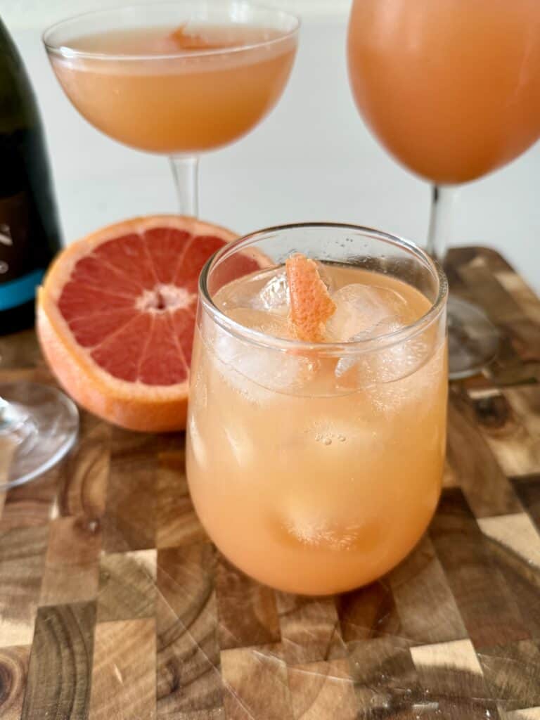 Grapefruit cocktail in a glass with ice, garnished with a slice of grapefruit, on a wooden surface with whole and halved grapefruits in the background.