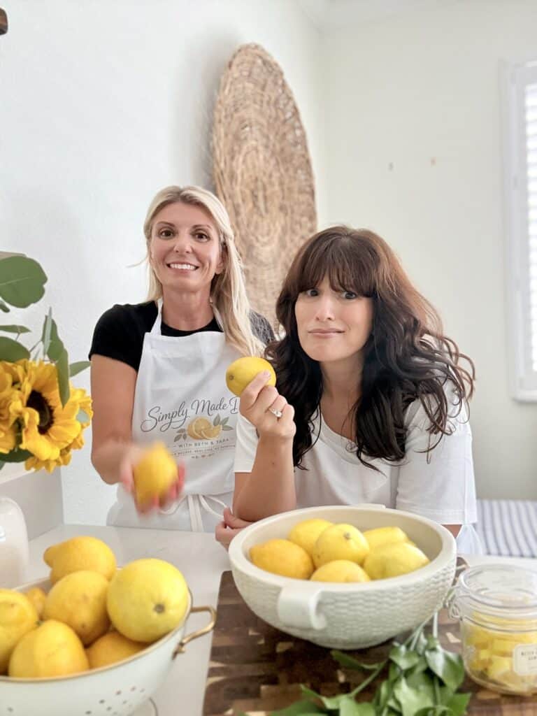 Two women in a bright kitchen preparing fresh lemons, one holding a lemon and the other smiling, with bowls of lemons on the table, showcasing healthy cooking.