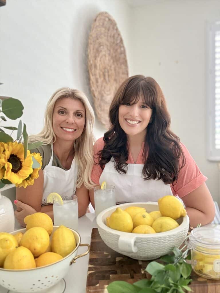 Two women, Beth and Tara, smiling in a bright kitchen with lemons, sunflowers, and homemade lemonade, showcasing a friendly cooking or baking scene.