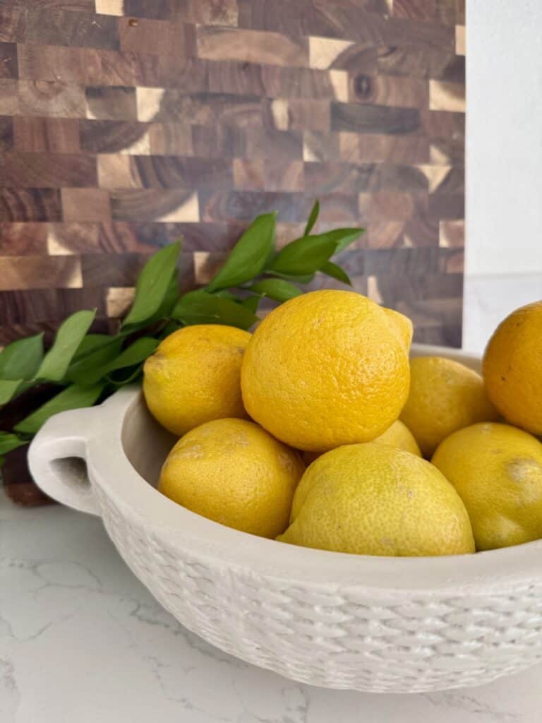 A white textured bowl filled with fresh, bright yellow lemons on a white marble surface with a wooden backsplash in the background.