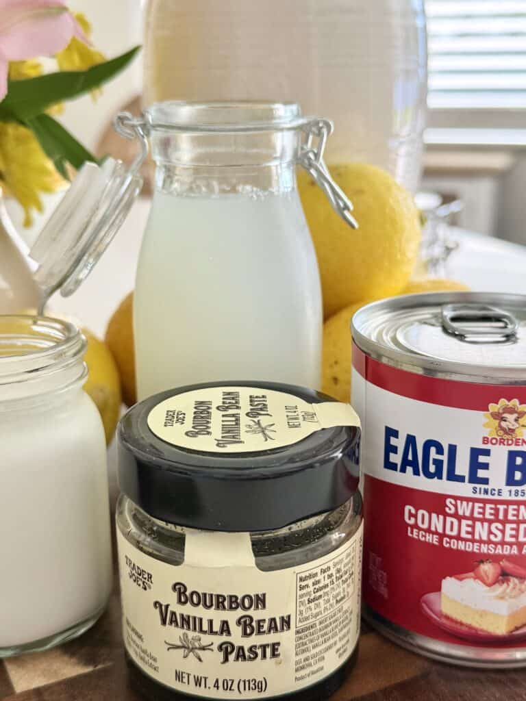 Lemonade and a clear glass behind it is lemonades and pink flowers and vases in a small clear jar is heavy, whipping cream, vanilla bean paste and sweetened condensed milk and a red and white and blue labeled can on a cutting board that is brown